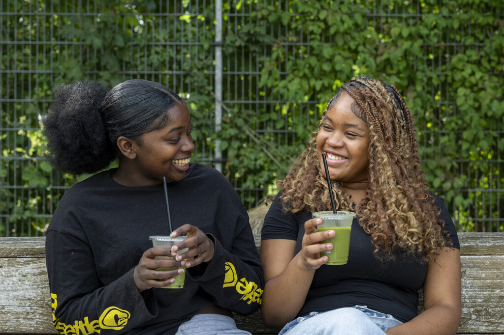 Twee vriendinnen van 15 en 16 zitten op een bankje met een beker Matcha in hun hand. Ze lachen naar elkaar.